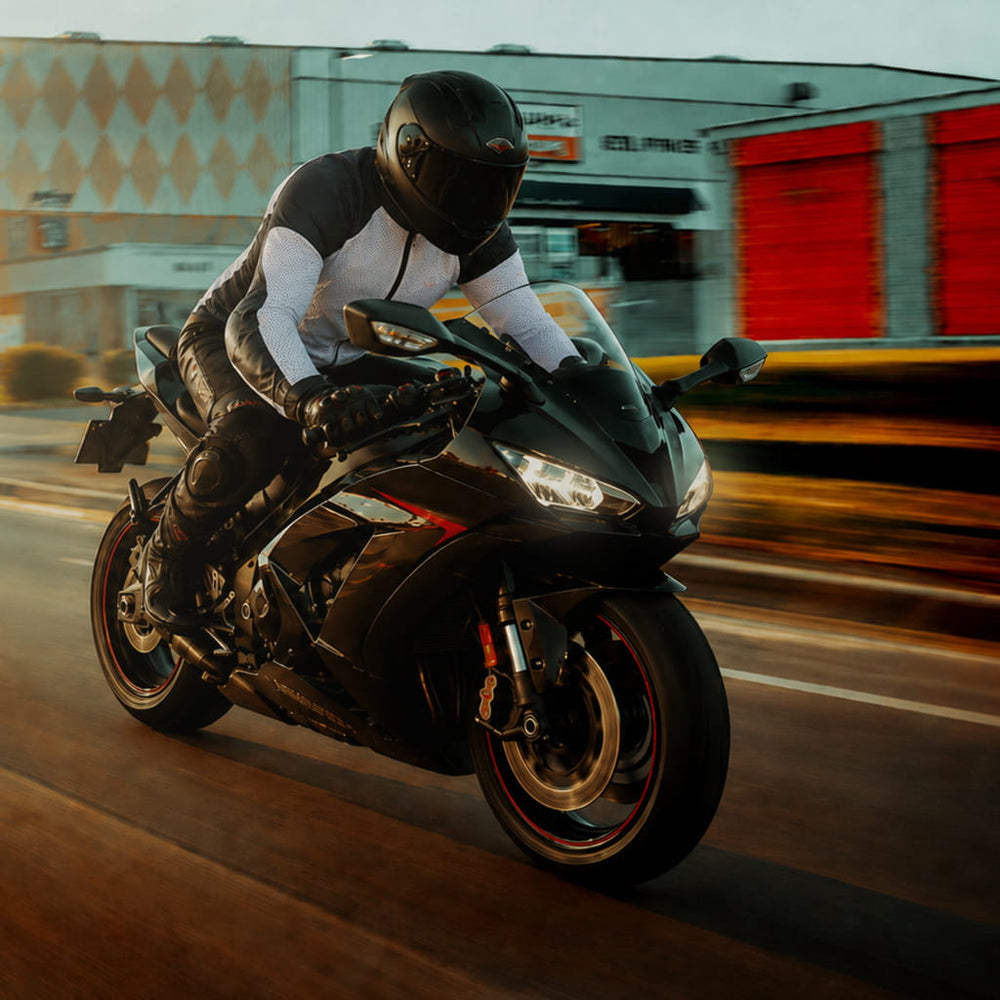 A person in a black, white, and gray Potomac Protective Riding Shirt Armored CE Level 2 Mesh All season riding a black sport motorcycle on a road with motion blur and a building in the background.
